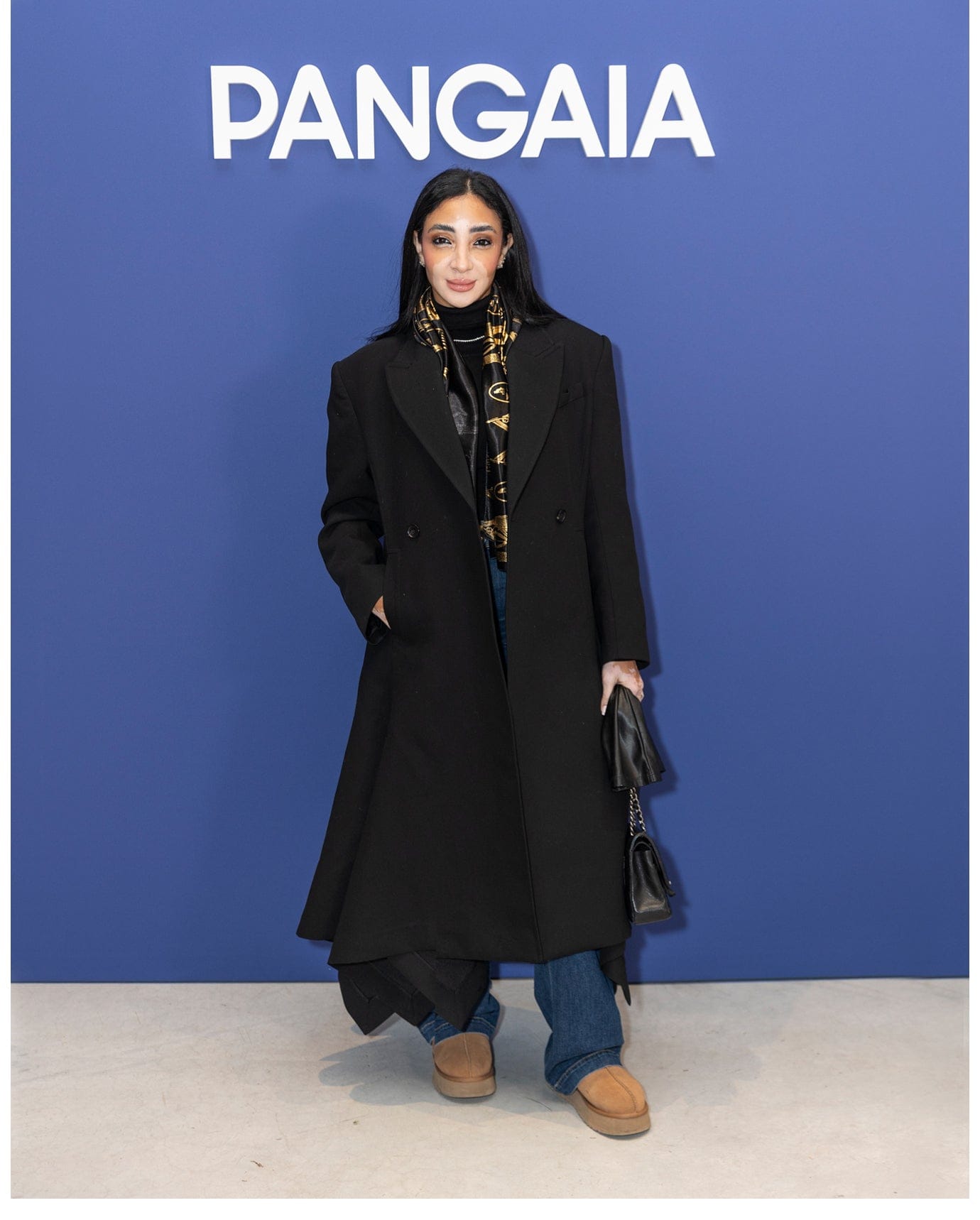 Woman wearing black structured wool coat, gold patterned scarf, black turtleneck, wide leg jeans, tan platform boots, black quilted bag