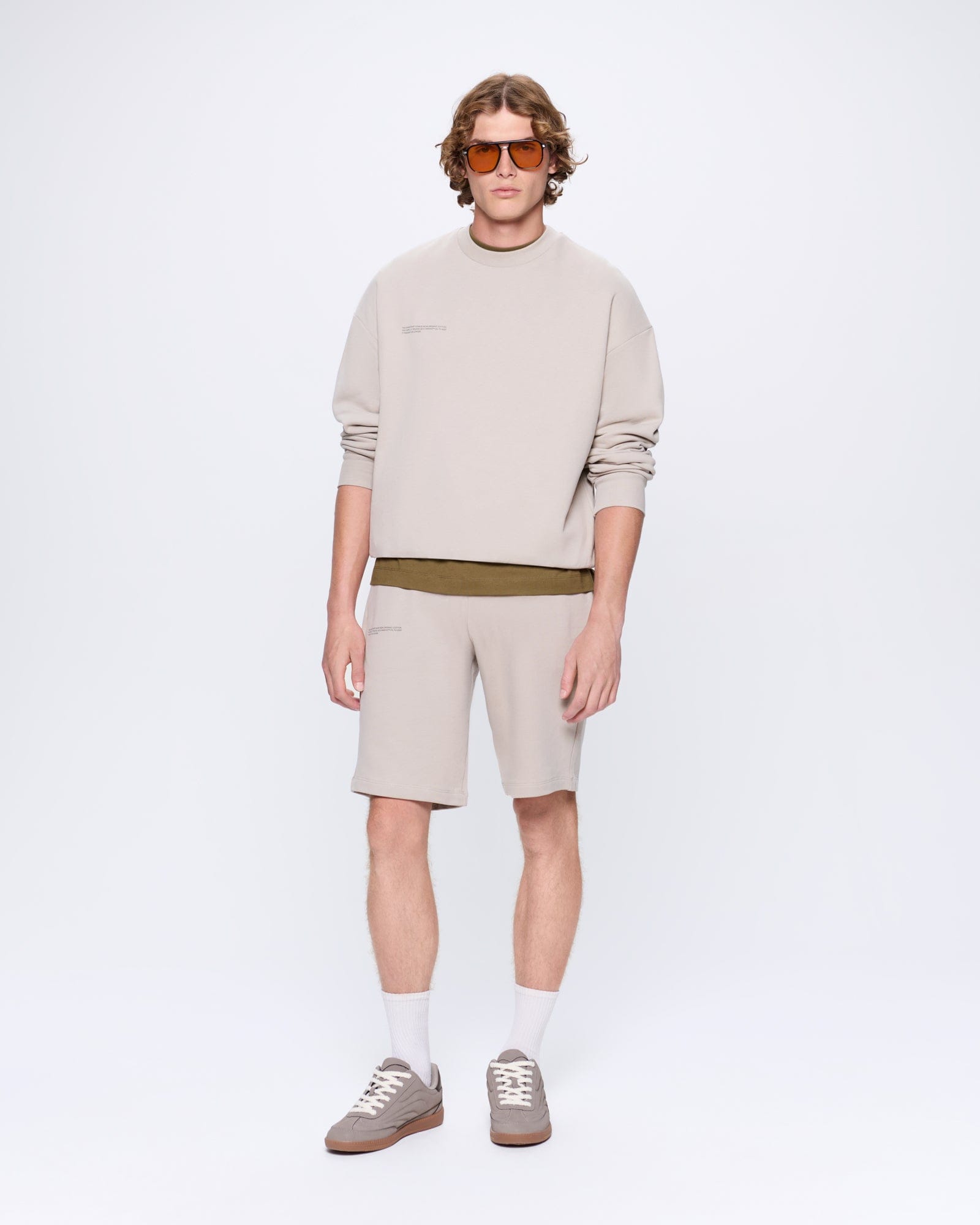 Men's relaxed fit greige crewneck sweatshirt & matching shorts. Olive tee, white socks, grey sneakers, orange sunglasses