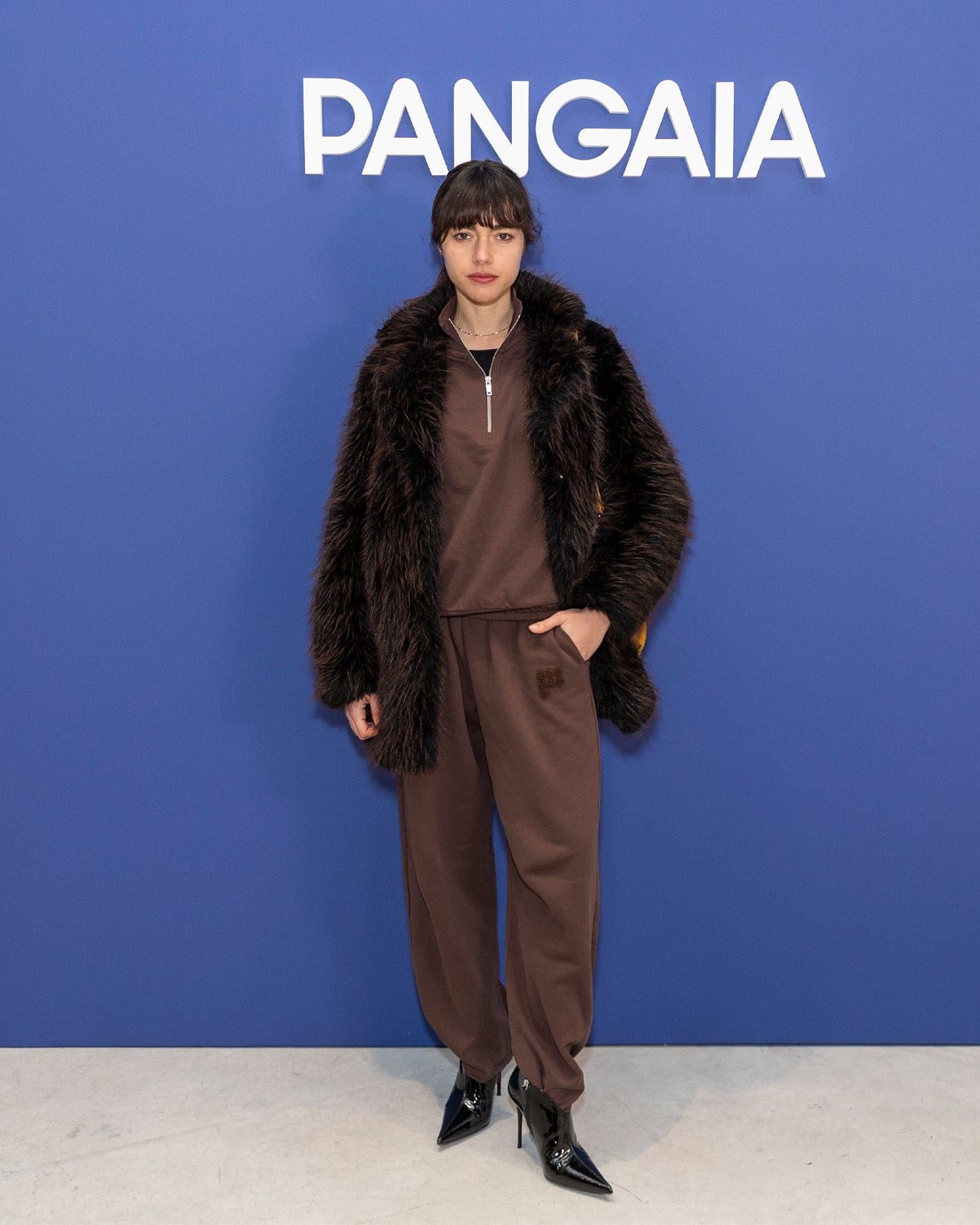 Model in Pangaia brown faux fur coat, brown quarter-zip sweatshirt, matching joggers, black pointed toe high heel boots