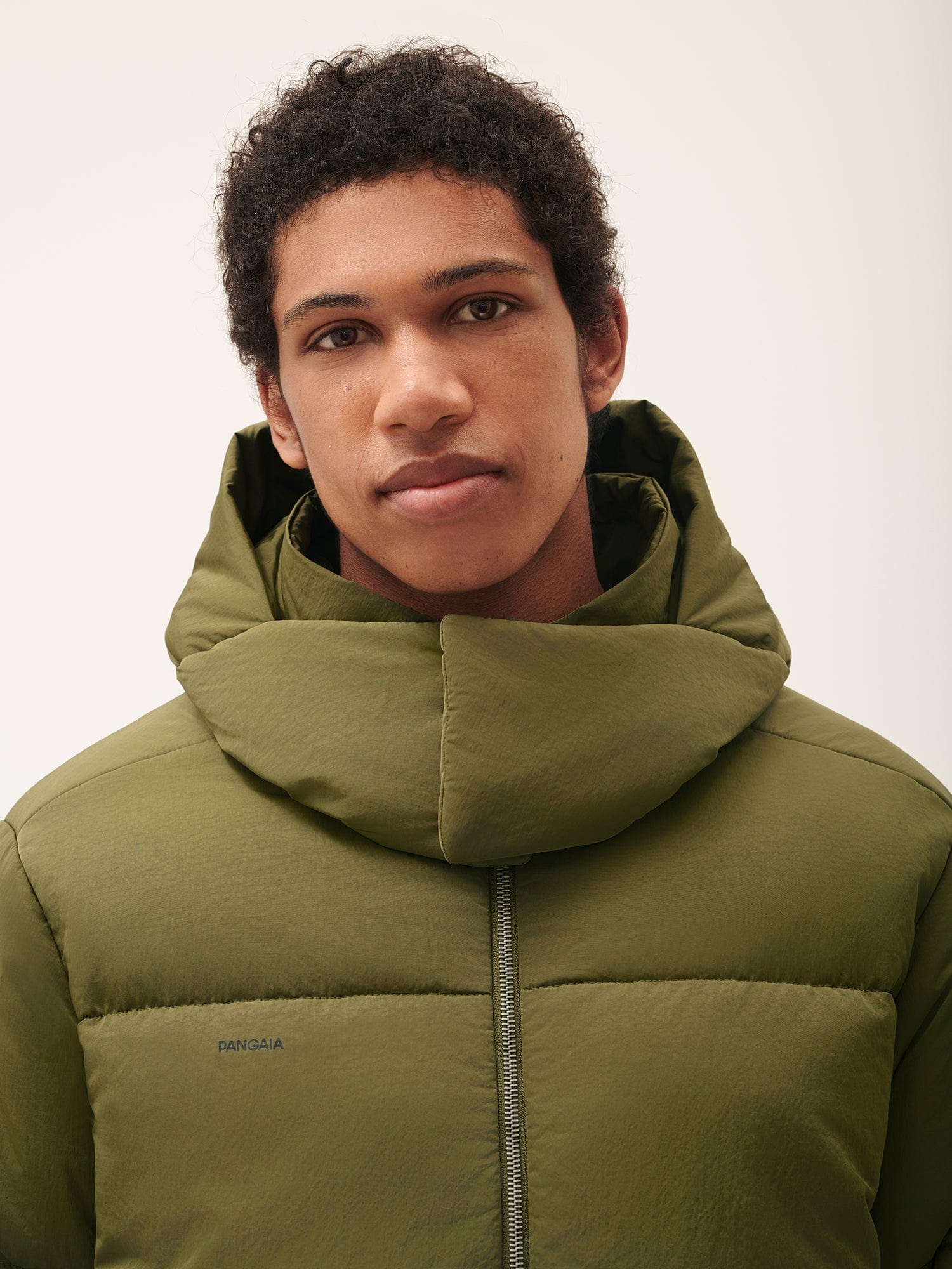 Pangaia men's olive green hooded puffer jacket. Model close-up wearing premium padded full-zip outerwear