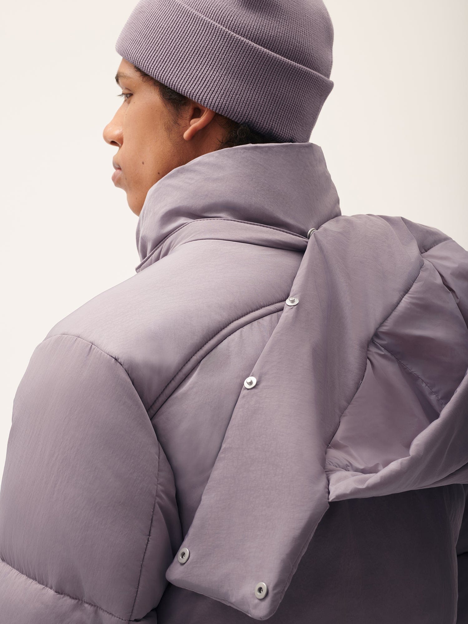 Men's lavender puffer jacket with high collar, hood snap detail, and matching ribbed knit beanie