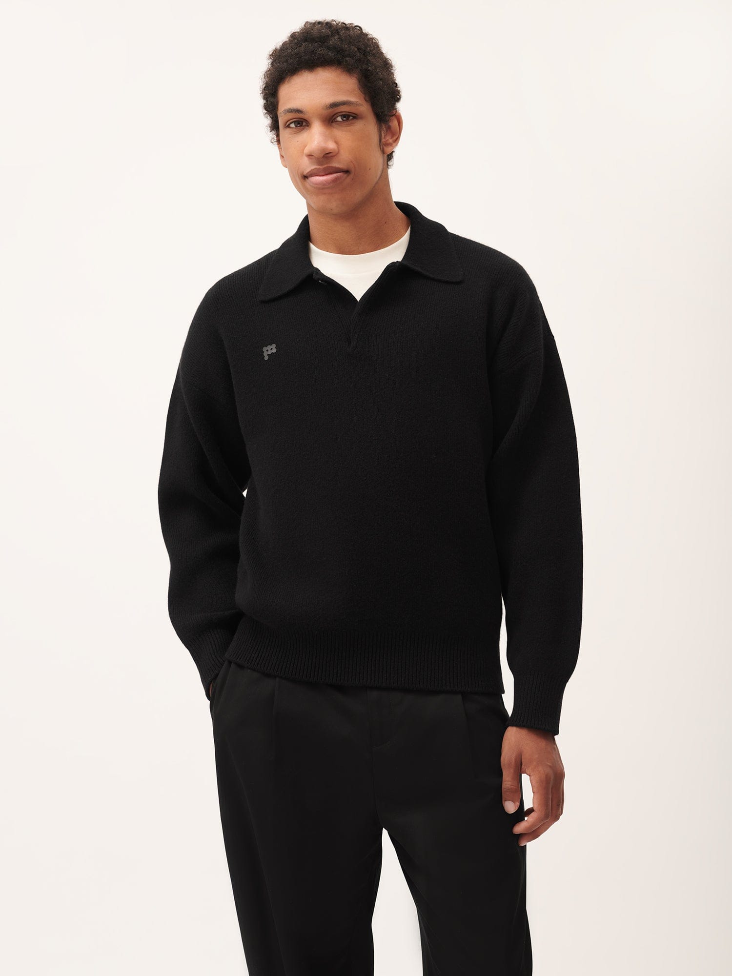Black fine knit men's polo sweater with spread collar ribbed cuffs subtle chest logo model wears black trousers