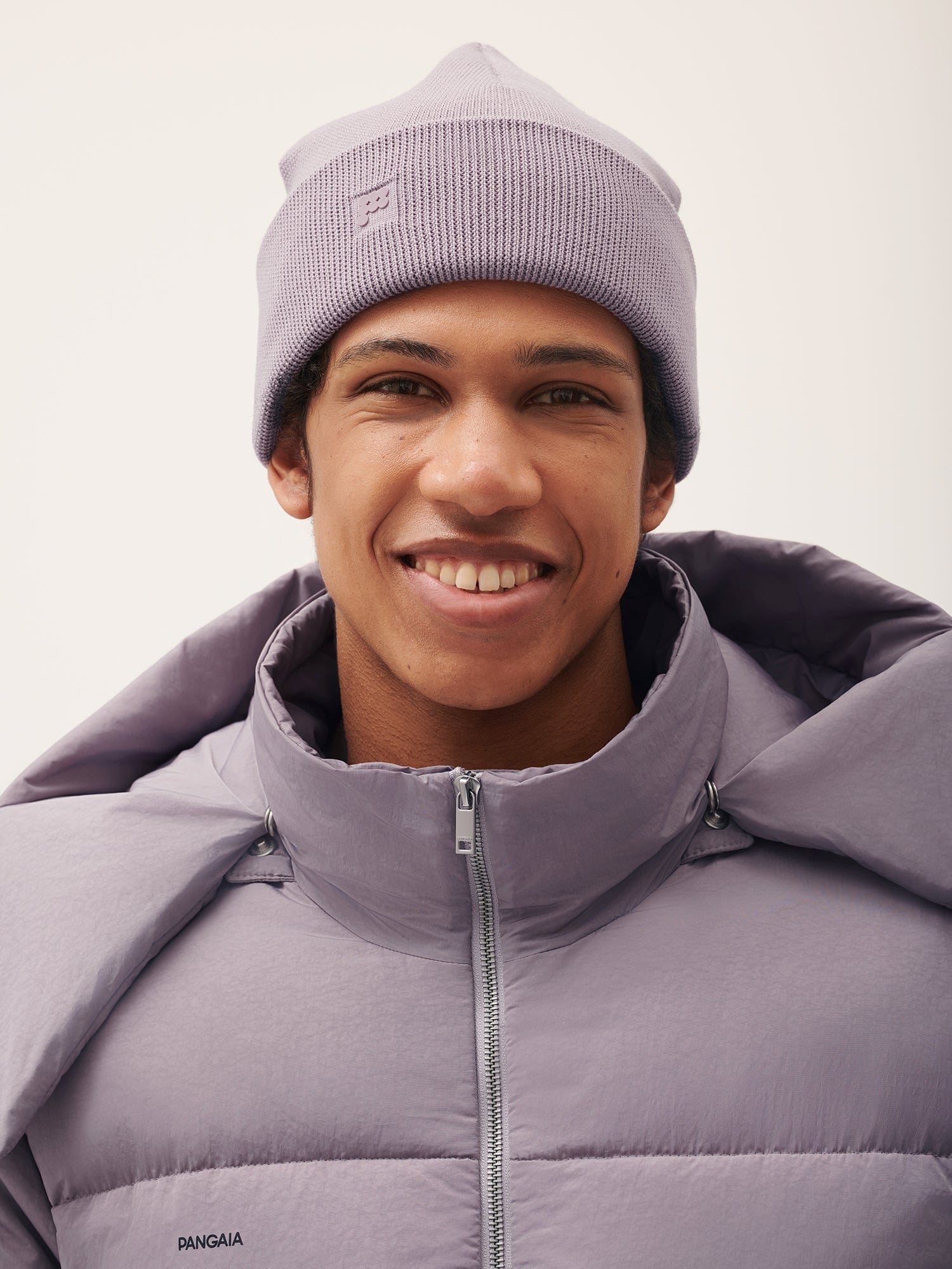 Smiling man in Pangaia lilac ribbed knit beanie with P logo, matching quilted puffer jacket, high zippered collar