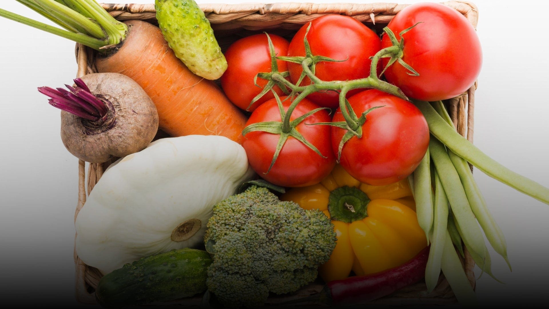 Wicker basket filled with diverse fresh vegetables: red tomatoes, carrots, broccoli, pepper, squash, beetroot, beans, cucumber