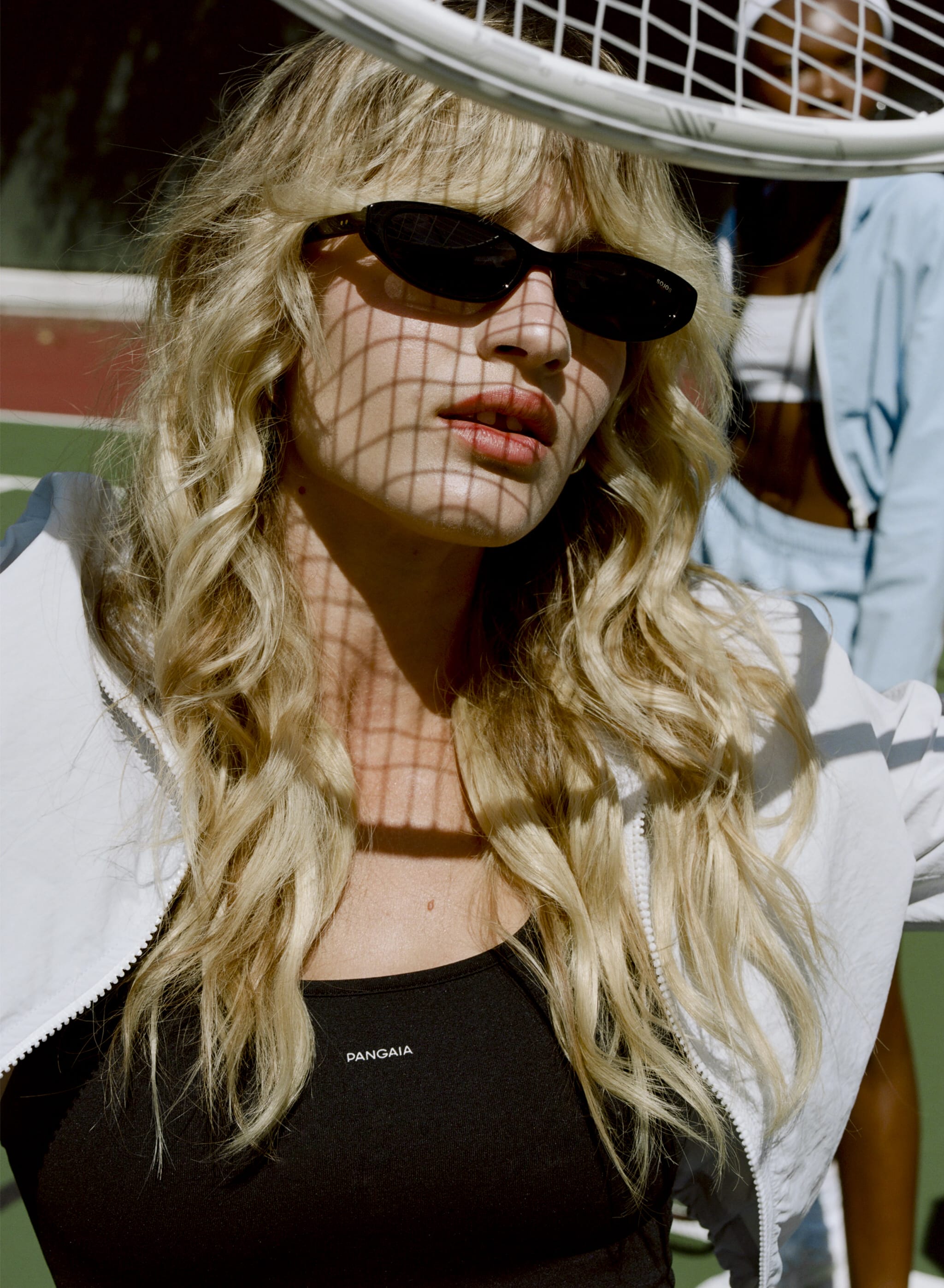 Blonde model wearing a black Pangaia fitted sports top and white crinkle nylon zip-up jacket for women's activewear Tennis court setting