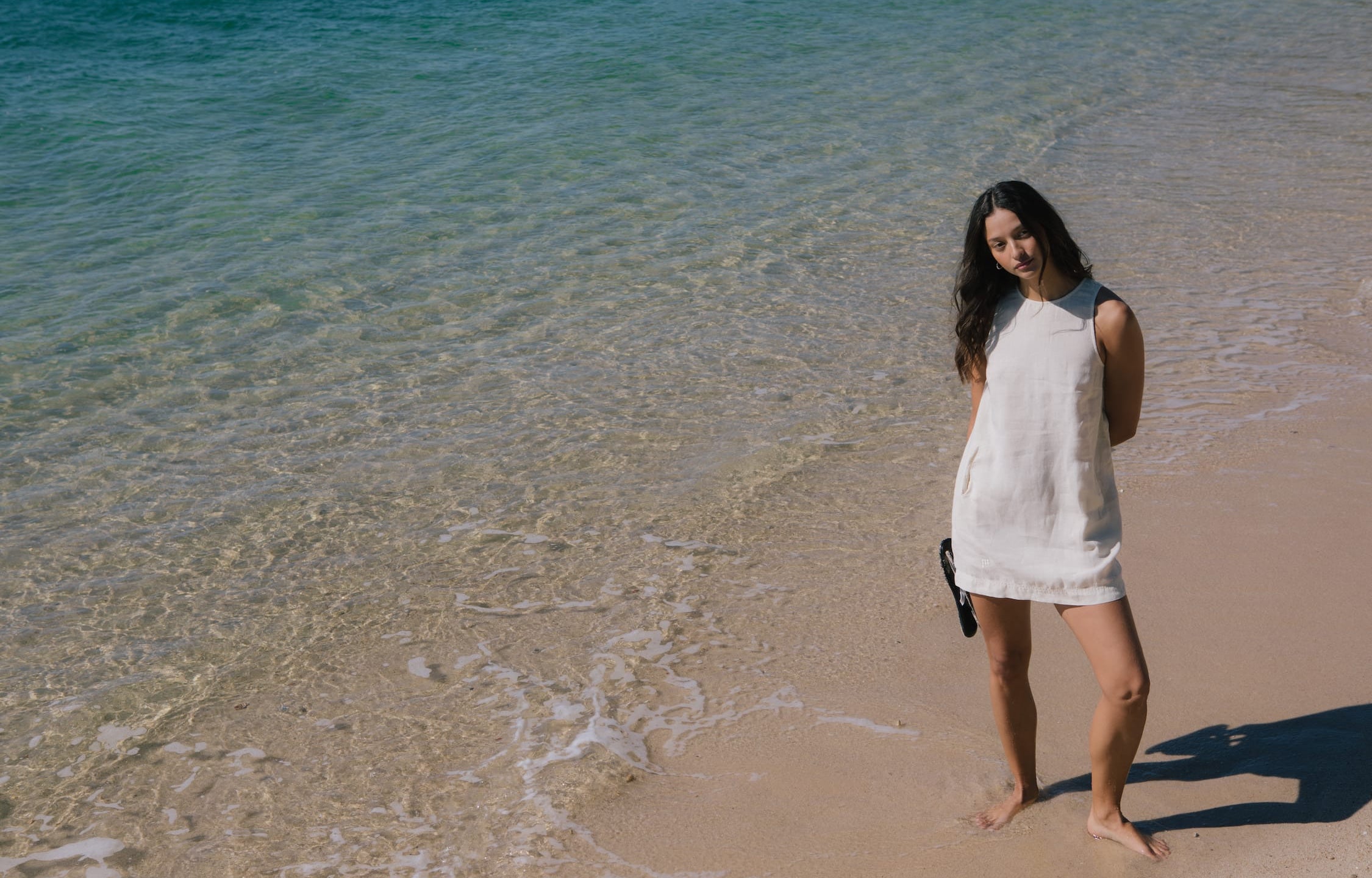 Model in cream linen blend sleeveless shift mini dress, relaxed fit, standing on sandy beach by clear ocean water