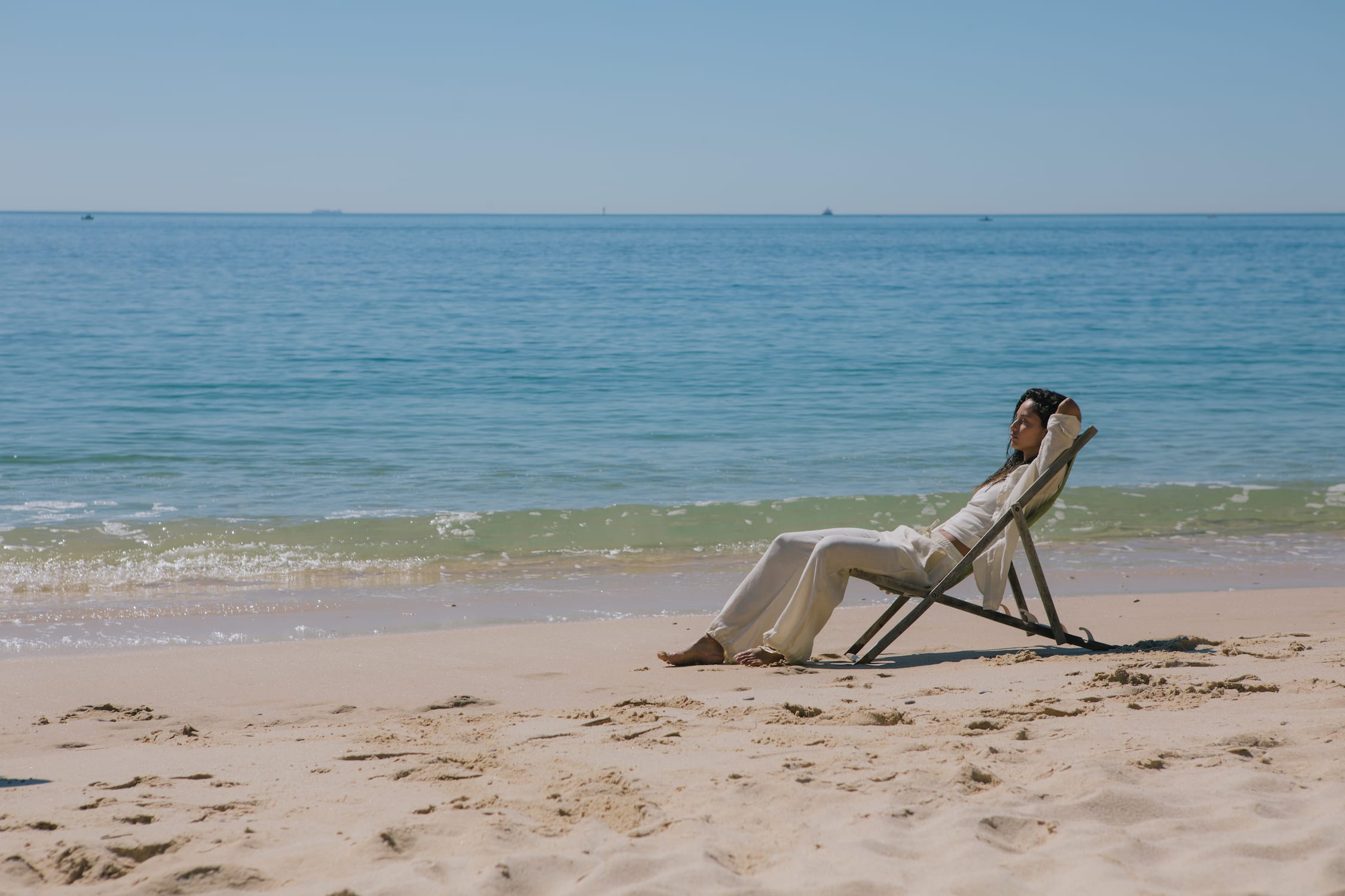 Woman wearing off-white linen blend long sleeve shirt and wide leg trousers, relaxed on beach. Premium resort wear.