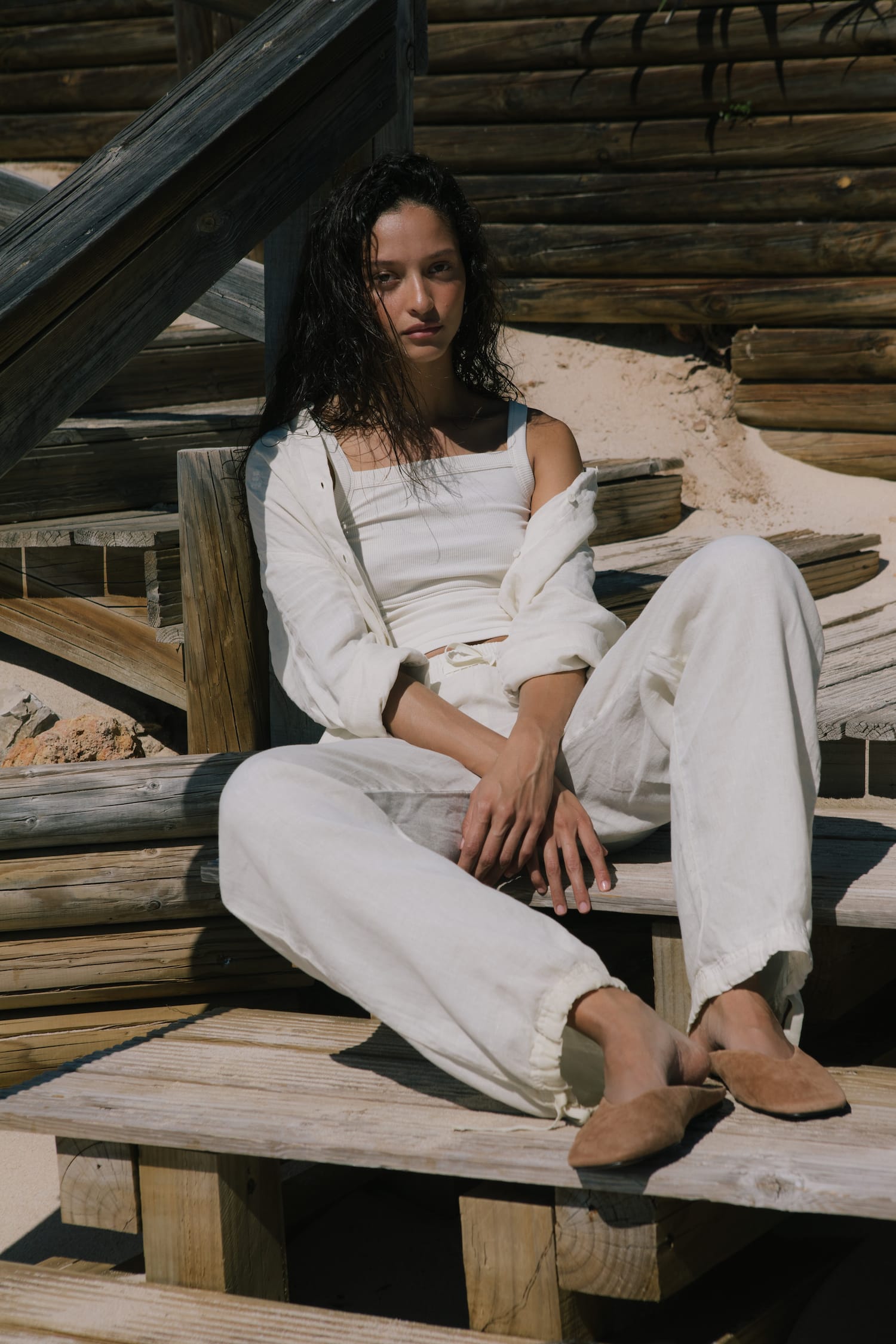 Model in off-white square neck ribbed tank, open linen shirt, relaxed fit linen trousers with drawstring cuffs, tan suede mules