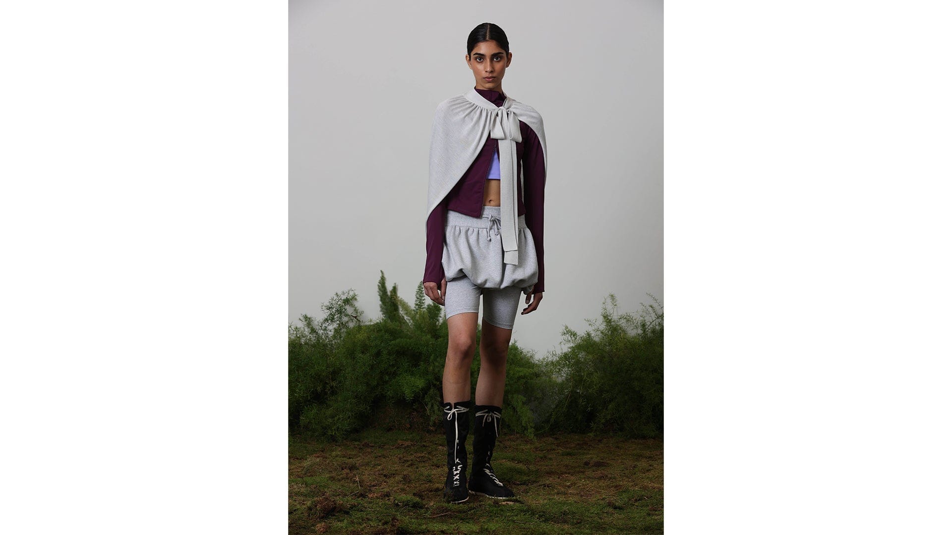 Model in deep purple long-sleeve, lavender crop, grey tied cape, slouchy shorts over biker shorts, black lace-up boots