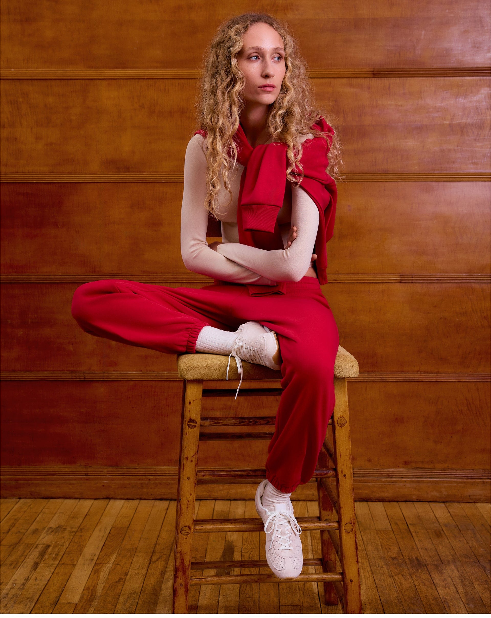 Blonde model wearing a red cotton fleece hoodie, matching red relaxed fit joggers, a beige long-sleeve top, and white sneakers