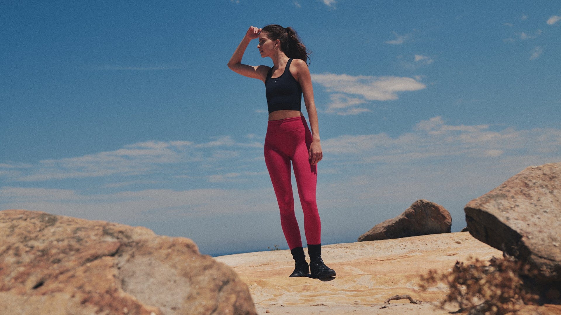 Woman wearing black racerback sports crop top and fuchsia high-waisted slim fit leggings against a rocky outdoor backdrop